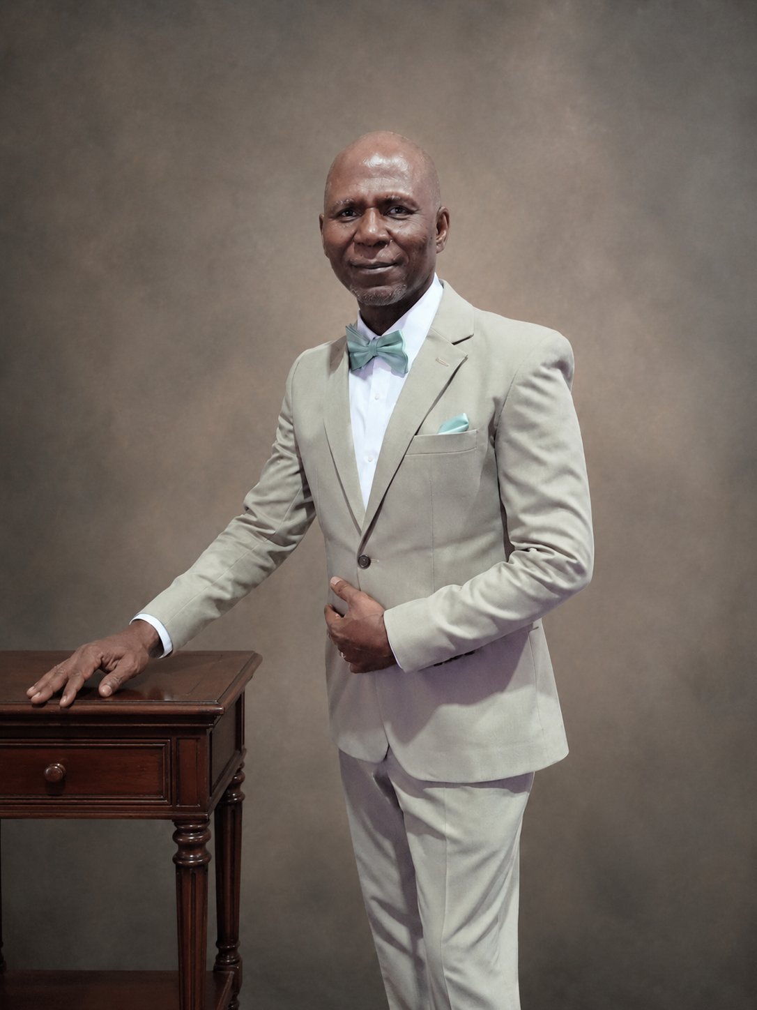 Professional portrait of a man in a cream-colored suit with blue bow tie, posing with hand on wooden table against neutral background